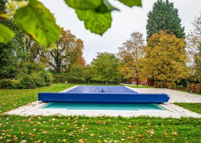 Une piscine couverte d'une bâche bleue est entourée de pelouse et d'arbres aux feuilles automnales dans un jardin.