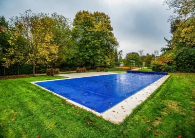 Piscine couverte d'une bâche bleue, entourée d'une pelouse verte et d'arbres aux feuilles automnales.