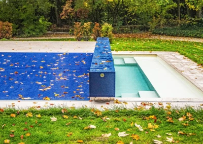 Une piscine partiellement couverte d'une bâche bleue, entourée de pelouse et d'arbres, avec des feuilles d'automne dispersées.