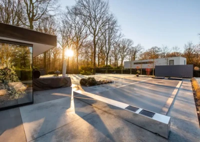 Terrasse moderne en béton avec plantes, entourée d'arbres dénudés et d'une maison blanche, éclairée par le soleil couchant.