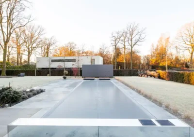 Piscine moderne recouverte d'une bâche grise dans un jardin avec pelouse, entourée d'arbres dénudés et d'une maison blanche en arrière-plan.