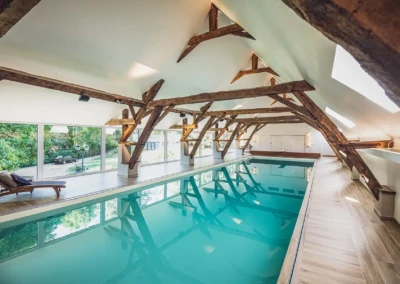 Piscine intérieure avec eau turquoise, entourée de poutres en bois et de fenêtres donnant sur un jardin verdoyant.
