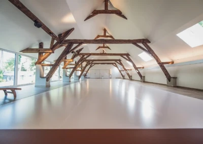 Grande salle avec poutres apparentes en bois sombre, plafond blanc incliné, fenêtres à gauche et puits de lumière à droite.