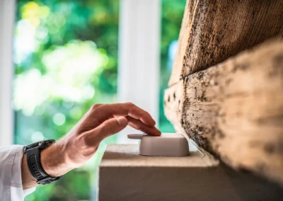 Une main appuie sur un interrupteur blanc fixé sur une poutre en bois, avec un fond flou de verdure.