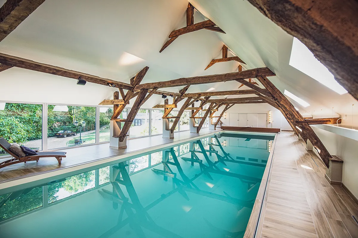 Piscine intérieure avec eau turquoise entourée de poutres en bois apparentes, fenêtres panoramiques et transat en bois.