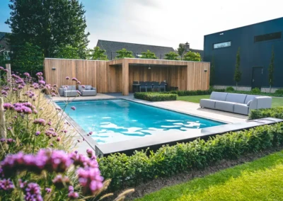 Piscine entourée de haies avec terrasse en bois, canapés gris et fleurs violettes au premier plan, maison moderne en arrière-plan.
