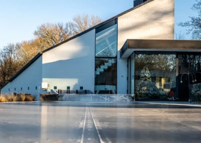 Maison moderne avec façade blanche et grandes fenêtres, entourée d'arbres dénudés, vue d'une large terrasse en béton.