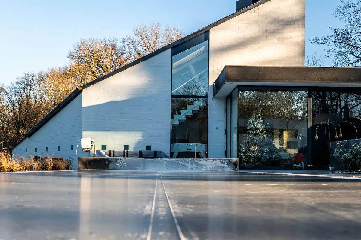 Maison moderne avec façade blanche et grandes fenêtres, entourée d'arbres dénudés, vue d'une large terrasse en béton.