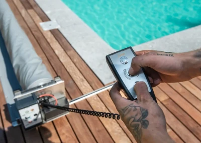 Une main tient une télécommande devant une piscine turquoise, avec un enrouleur de bâche sur une terrasse en bois.