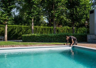 Un homme installe un dispositif au bord d'une piscine entourée d'arbres et de haies, sous un ciel dégagé.