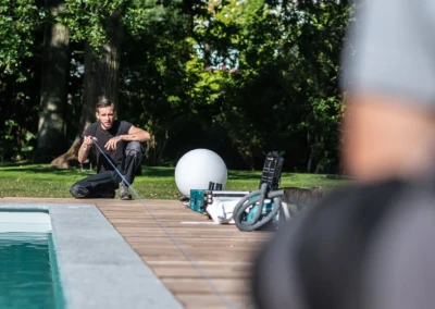 Un homme agenouillé près d'une piscine tient un ruban à mesurer, entouré d'outils et de verdure en arrière-plan.