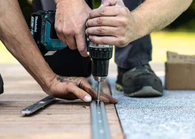 Deux mains utilisent une perceuse électrique pour fixer une vis sur une planche en bois, avec une boîte en carton à proximité.