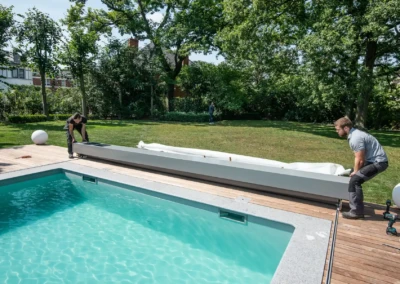 Deux personnes installent un dispositif de couverture sur une piscine entourée de bois, avec un jardin verdoyant en arrière-plan.