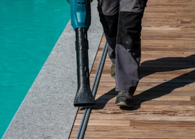 Une personne en pantalon gris marche sur un sol en bois près d'une piscine turquoise, tenant un équipement de nettoyage noir.