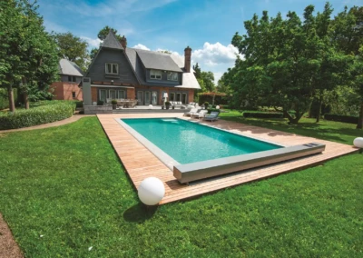 Maison en briques avec toit en ardoise, piscine rectangulaire entourée de terrasse en bois, pelouse verte et arbres environnants.