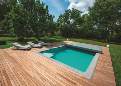 Quatre chaises longues grises sont disposées sur une terrasse en bois autour d'une piscine rectangulaire dans un jardin verdoyant.