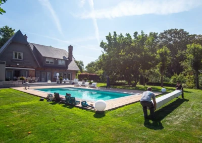 Deux personnes installent un dispositif blanc autour d'une piscine entourée de chaises longues, devant une maison en briques avec jardin.