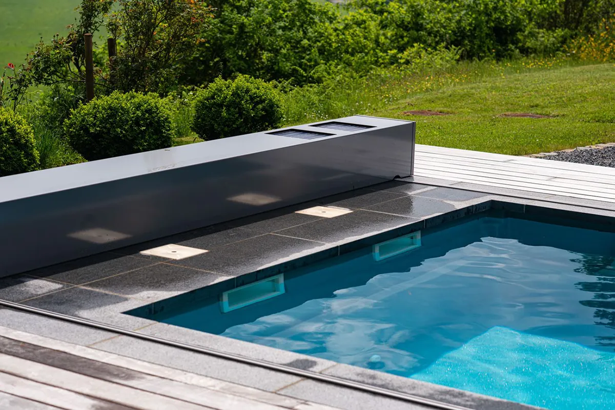 Piscine avec margelles en pierre grise, entourée de verdure et d'une terrasse en bois clair.