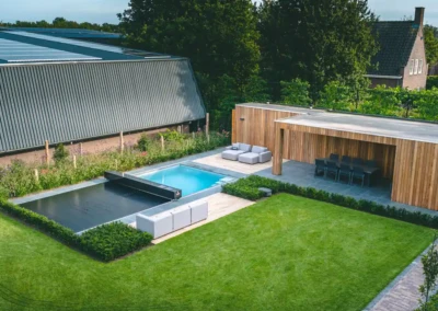 Piscine extérieure couverte à côté d'un salon de jardin en bois avec des canapés gris, entourée de pelouse et de haies.