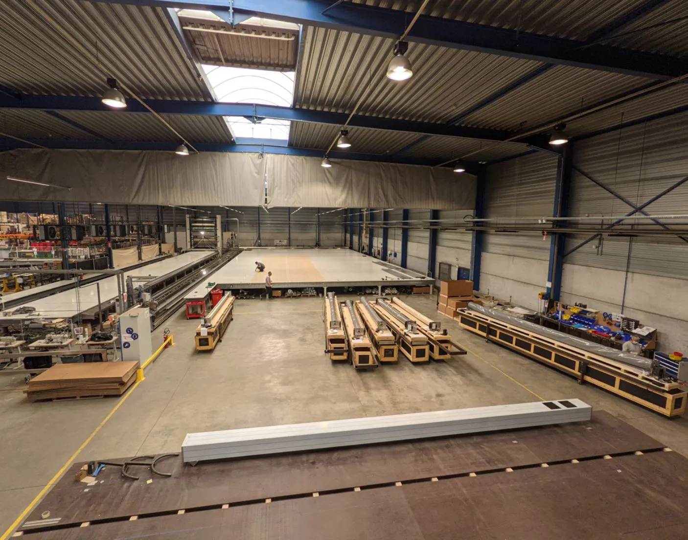 COVERSEAL-entreprise-4 Une grande usine avec des machines industrielles et des matériaux en bois disposés au centre, sous un toit métallique avec des lumières suspendues.