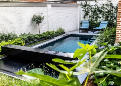 Une piscine rectangulaire entourée de plantes vertes et de murs en briques blanches, avec deux chaises longues bleues posées sur une terrasse grise.