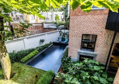 Une piscine couverte noire est entourée de verdure, avec deux chaises longues bleues, près d'une maison en briques rouges.
