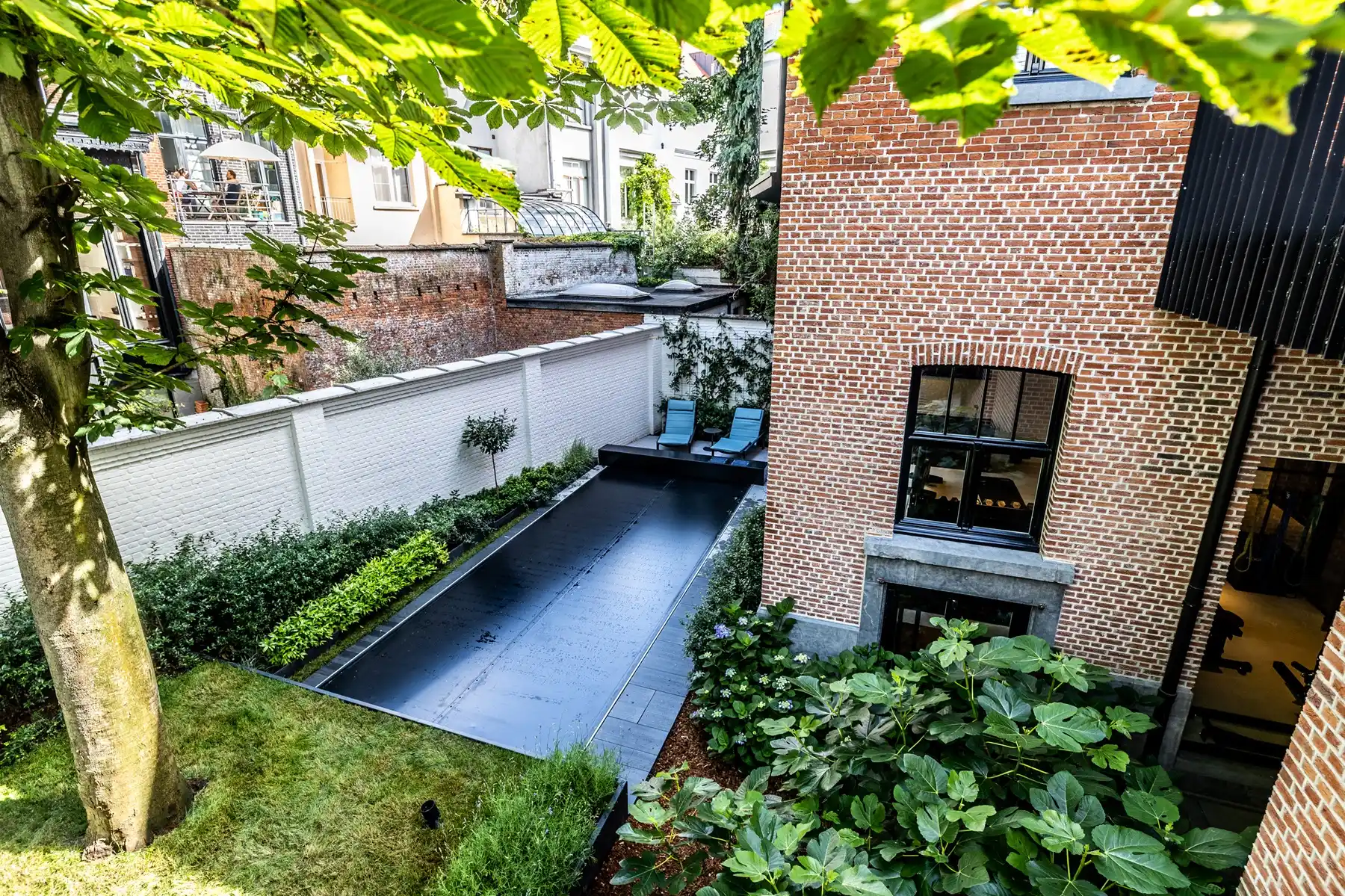 Chez-Anna-et-Luc-3 Une piscine couverte noire est entourée de verdure, avec deux chaises longues bleues, près d'une maison en briques rouges.