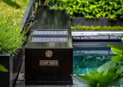 Un boîtier noir avec panneaux solaires est installé près d'une piscine, entouré de plantes vertes.