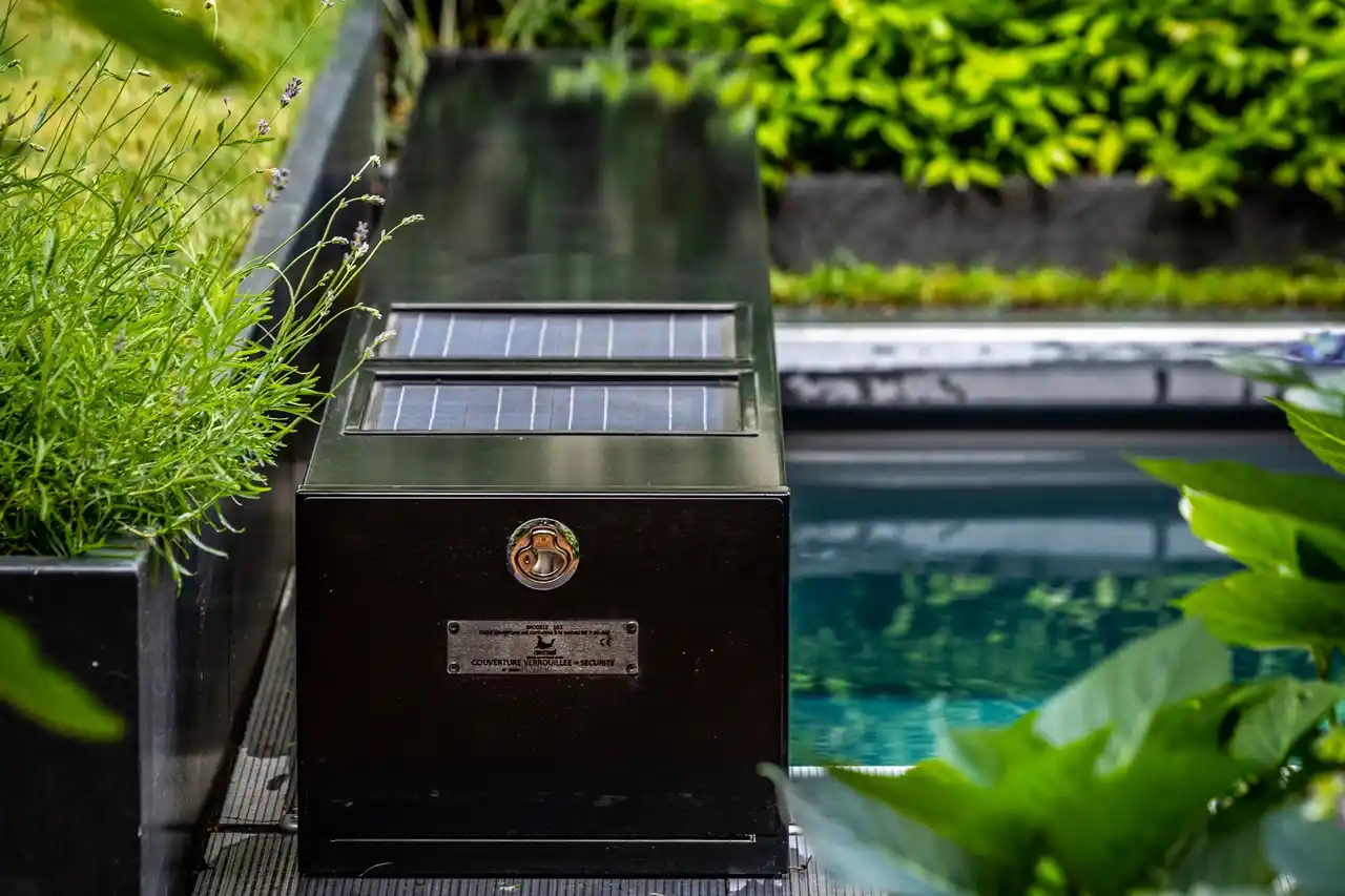 Chez-Anna-et-Luc-4 Un boîtier noir avec panneaux solaires est installé près d'une piscine, entouré de plantes vertes.