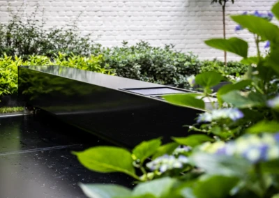 Une table noire rectangulaire est placée au milieu d'un jardin avec des buissons verts et un mur de briques blanches en arrière-plan.