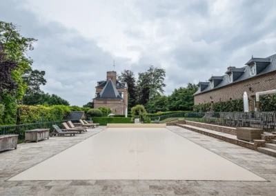 Terrasse en pierre avec piscine couverte, entourée de chaises longues en bois et d'une maison en briques sous un ciel nuageux.
