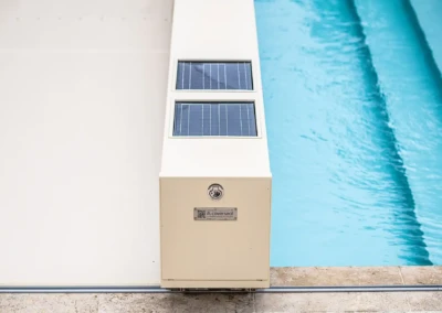 Un boîtier beige avec panneaux solaires est installé au bord d'une piscine bleue, sur un sol en pierre.