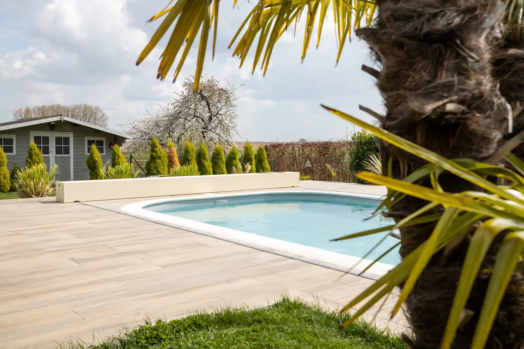 Chez-Charlotte-et-Pierre-1 Une piscine entourée de dalles en bois clair avec un cabanon vert et des arbres en arrière-plan sous un ciel nuageux.
