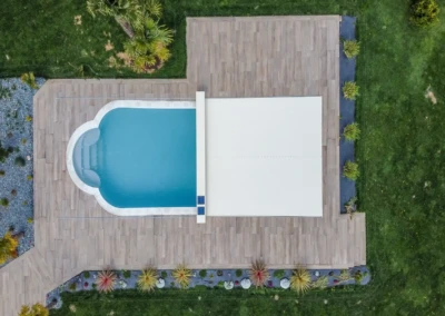 Vue aérienne d'une piscine rectangulaire avec une couverture blanche, entourée de dalles en bois et de pelouse verte.
