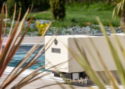 Un boîtier beige de piscine Coverseal près de l'eau, entouré de végétation et d'un jardin verdoyant.
