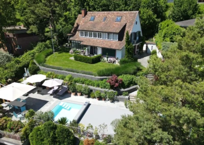 Maison avec toit en tuiles entourée de jardins verdoyants, piscine, et parasols blancs sur terrasse ensoleillée.