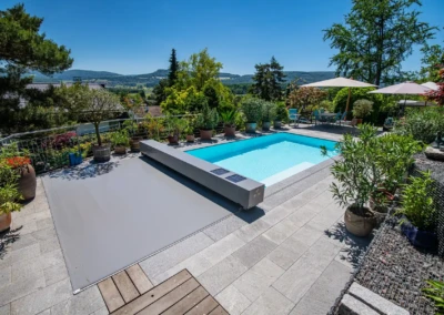 Piscine rectangulaire entourée de plantes en pots, avec parasols et paysage de collines en arrière-plan sous un ciel bleu.