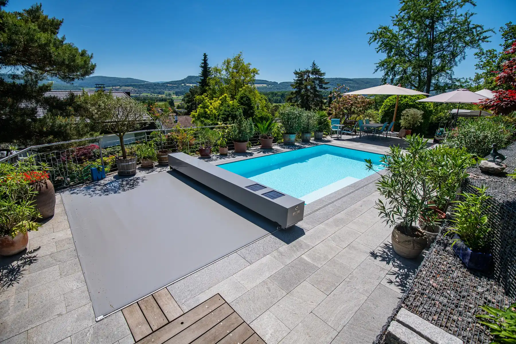 Piscine rectangulaire entourée de plantes en pots, avec parasols et paysage de collines en arrière-plan sous un ciel bleu.