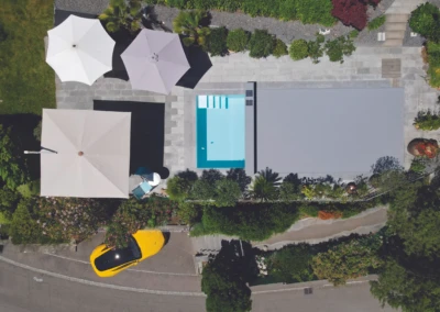 Vue aérienne d'une piscine rectangulaire entourée de parasols blancs et beige, avec une voiture jaune garée à proximité.