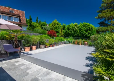 Piscine couverte de gris entourée de plantes en pots, avec un parasol et une chaise longue sous un ciel bleu.
