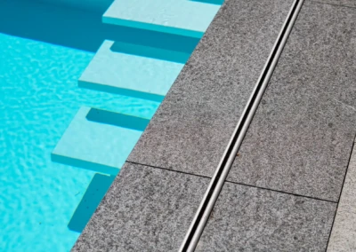 Dalles en pierre grise bordant une piscine avec des marches immergées dans l'eau turquoise.
