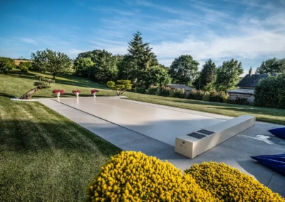 Jardin avec pelouse verte, piscine couverte, pots de fleurs roses, arbustes et arbres en arrière-plan sous un ciel bleu.