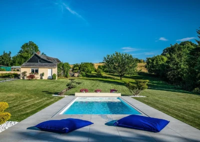 Piscine rectangulaire entourée de coussins bleus, située dans un jardin avec pelouse, arbres et une maison beige au fond.