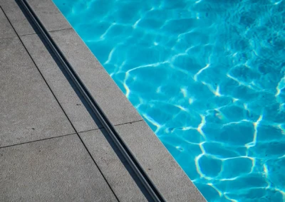 Bord de piscine en dalles grises avec eau bleue scintillante à droite, séparées par une fine rainure métallique.