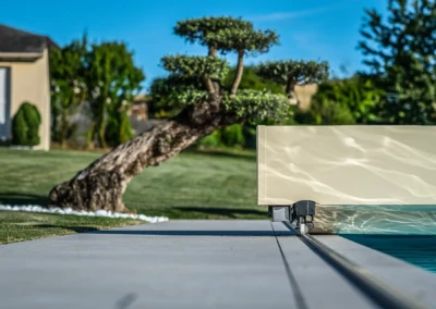 Couverture de piscine beige au bord d'une piscine, avec un arbre taillé en arrière-plan sur une pelouse verte.