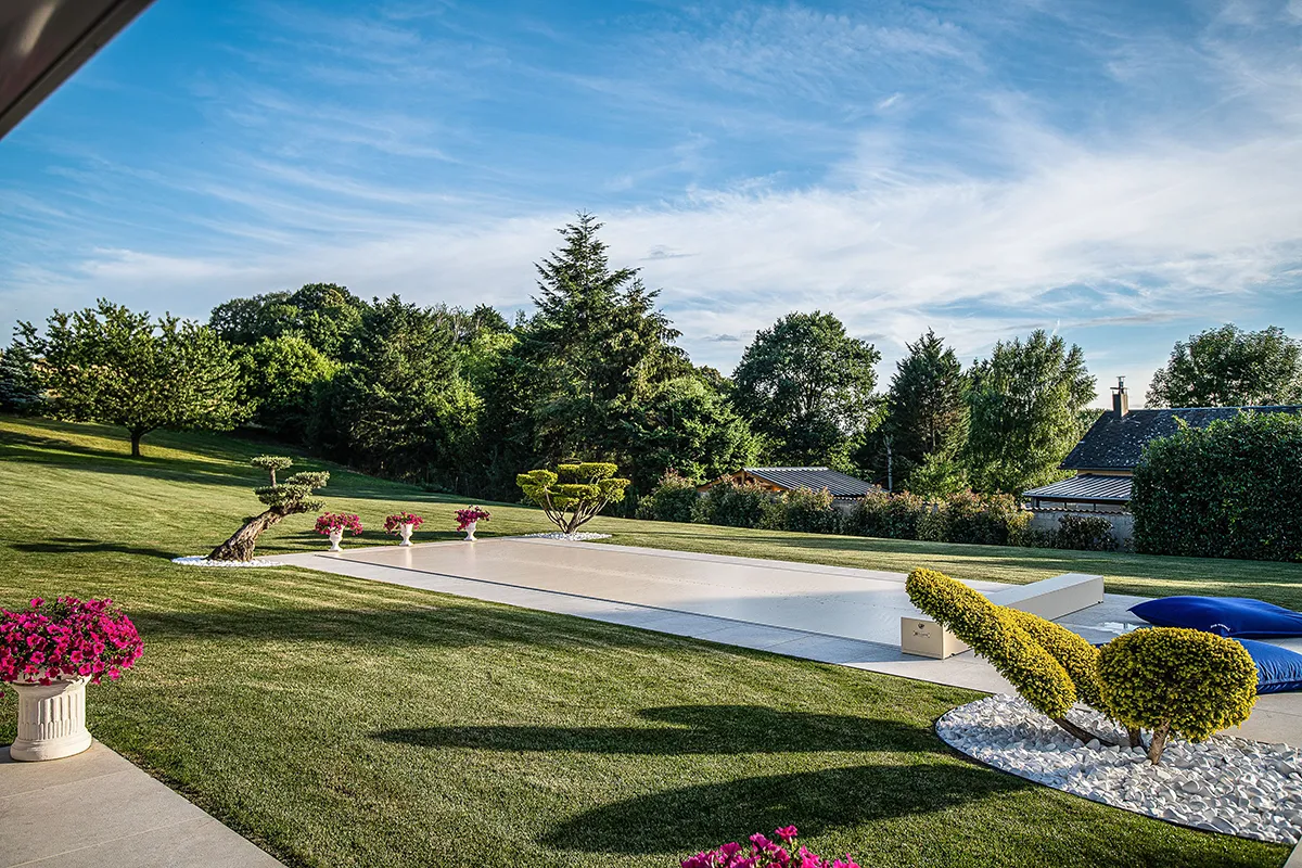Un jardin paysager avec une piscine couverte, entouré de pelouse verte, d'arbustes taillés et de pots de fleurs roses.
