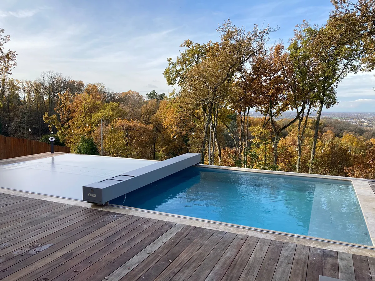 Piscine rectangulaire avec couverture grise sur terrasse en bois, entourée d'arbres aux feuilles automnales.
