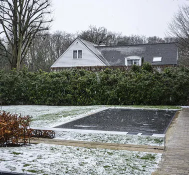 Maison grise entourée d'arbres, avec piscine couverte et jardin enneigé, bordée de haies et allée pavée.