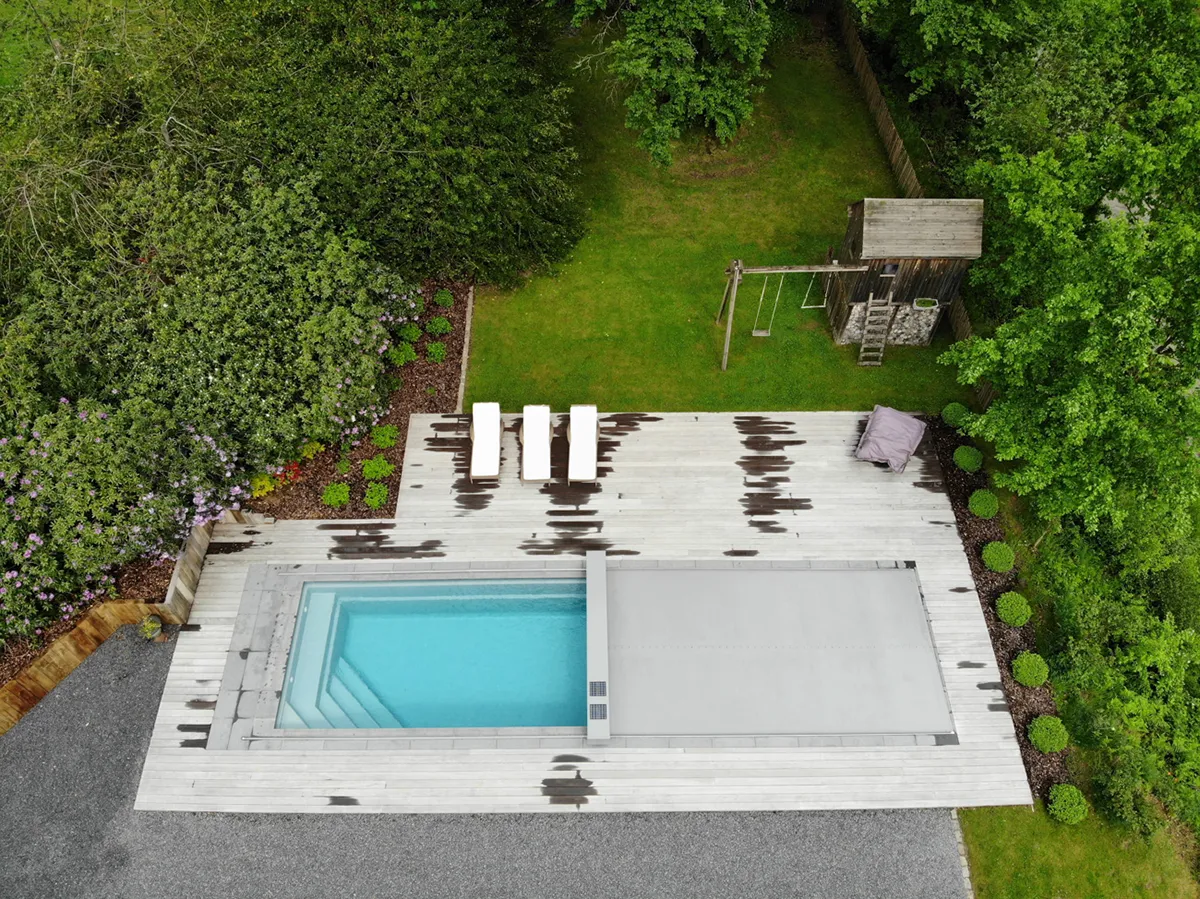 Vue aérienne d'une piscine rectangulaire avec couverture grise, entourée de chaises longues blanches sur une terrasse en bois, près d'un jardin verdoyant.