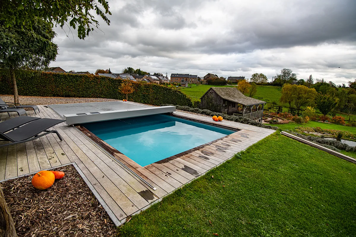 Piscine rectangulaire avec couverture grise sur terrasse en bois, entourée de pelouse verte et de citrouilles orange, paysage de campagne en arrière-plan.